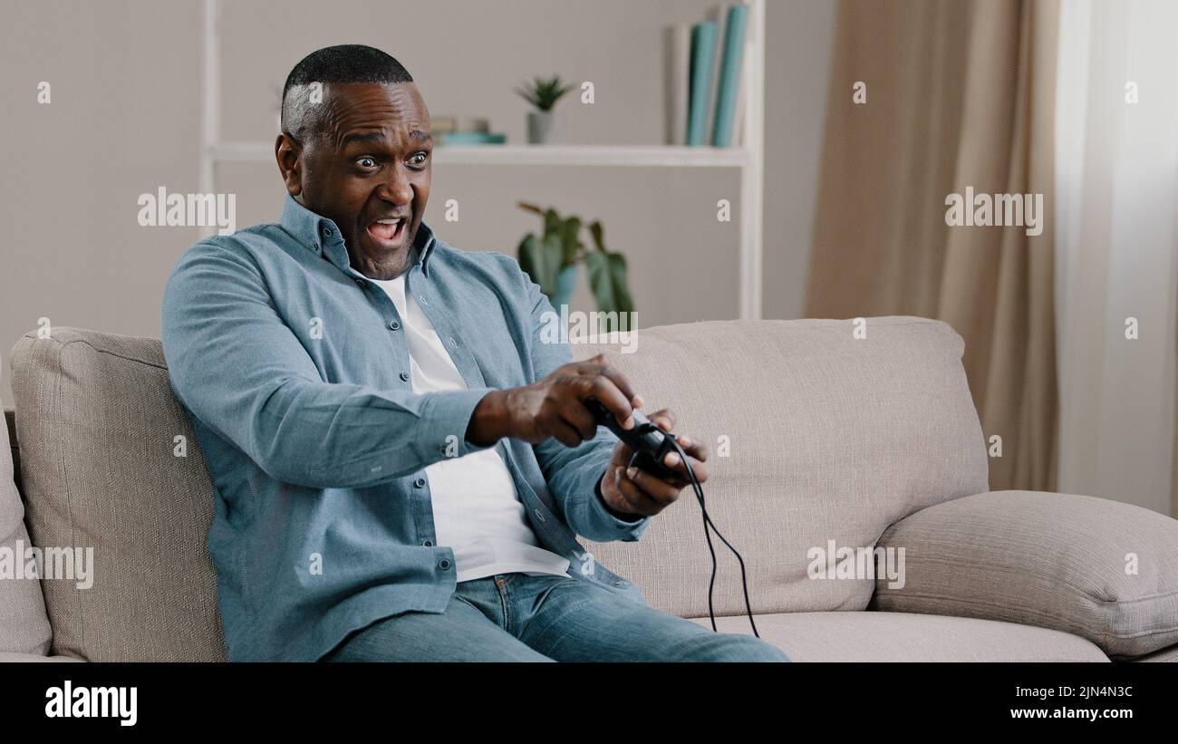 Aufgeregt besorgt emotionale Erwachsene afroamerikanische männliche Gamer auf dem Sofa im Wohnzimmer sitzend Videospiel genießt Hobby halten Controller Joystick spielen Stockfoto