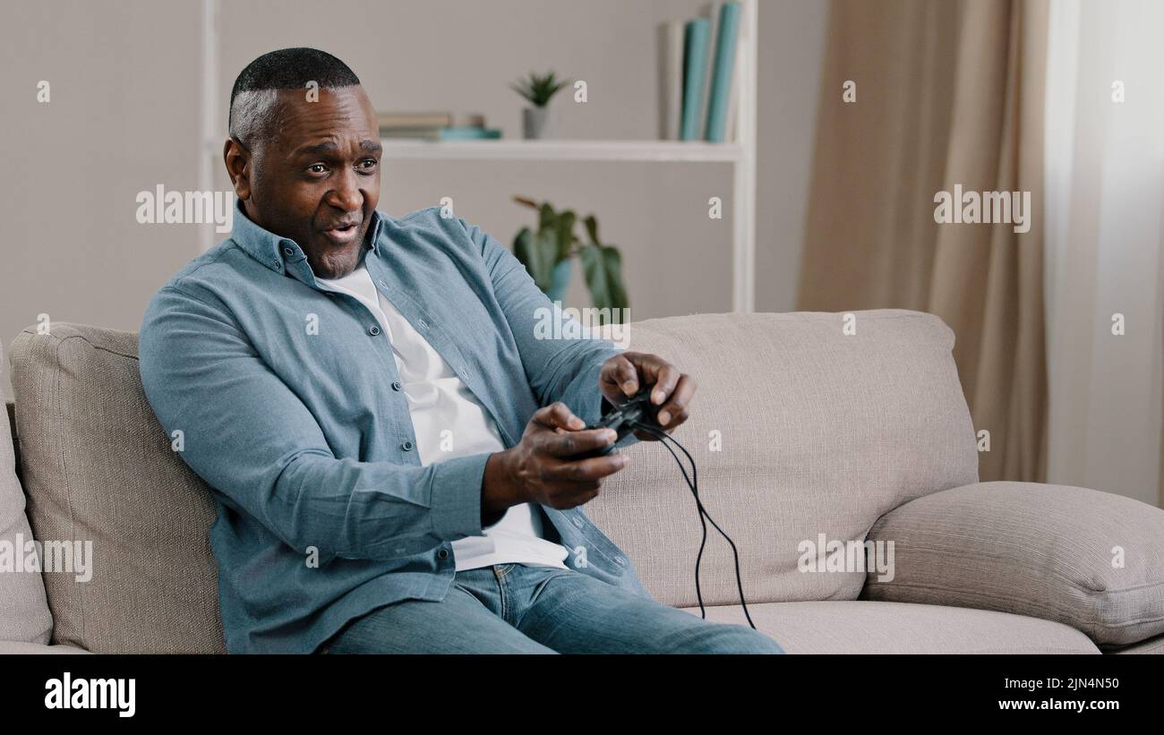Aufgeregt besorgt emotionale Erwachsene afroamerikanische männliche Gamer auf dem Sofa im Wohnzimmer sitzend Videospiel genießt Hobby halten Controller Joystick spielen Stockfoto