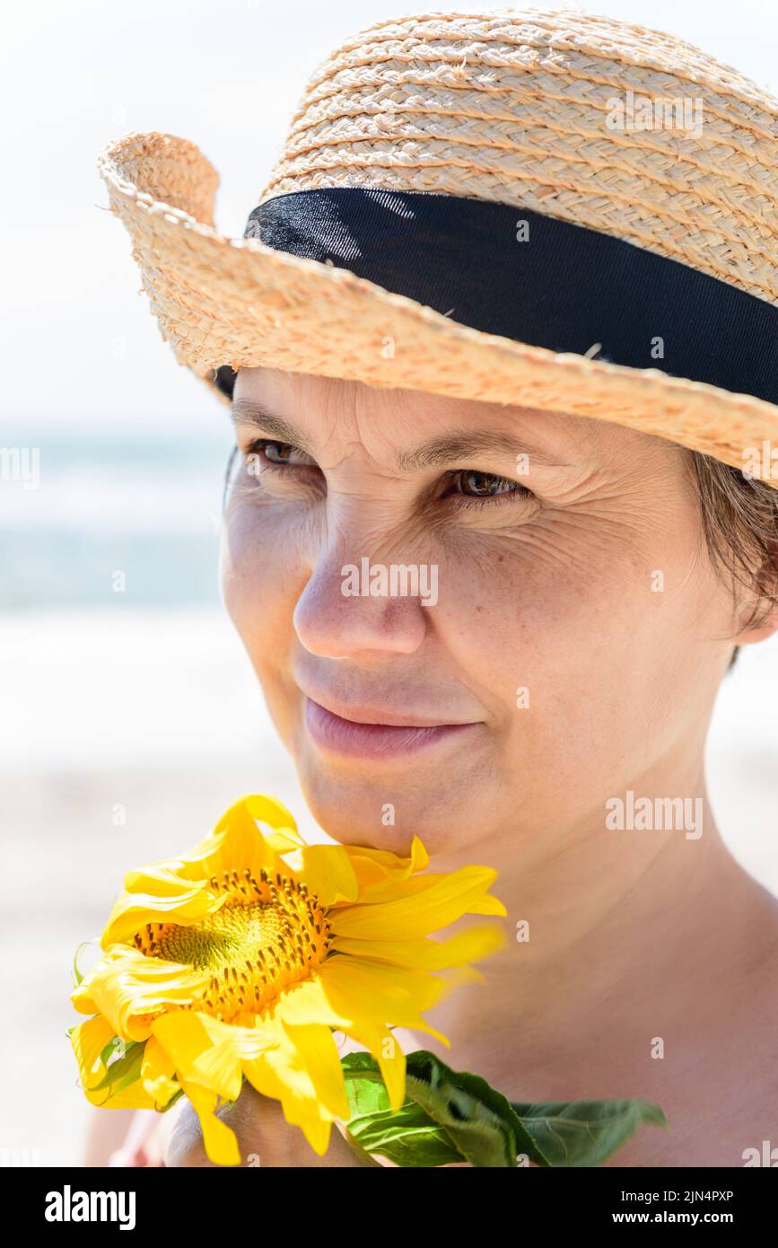 Frau in einem Strohhut mit Sonnenblume schaut zur Seite und wendet ihr Gesicht dem starken Wind, vor verschwommenem Hintergrund des Meeres. Stockfoto