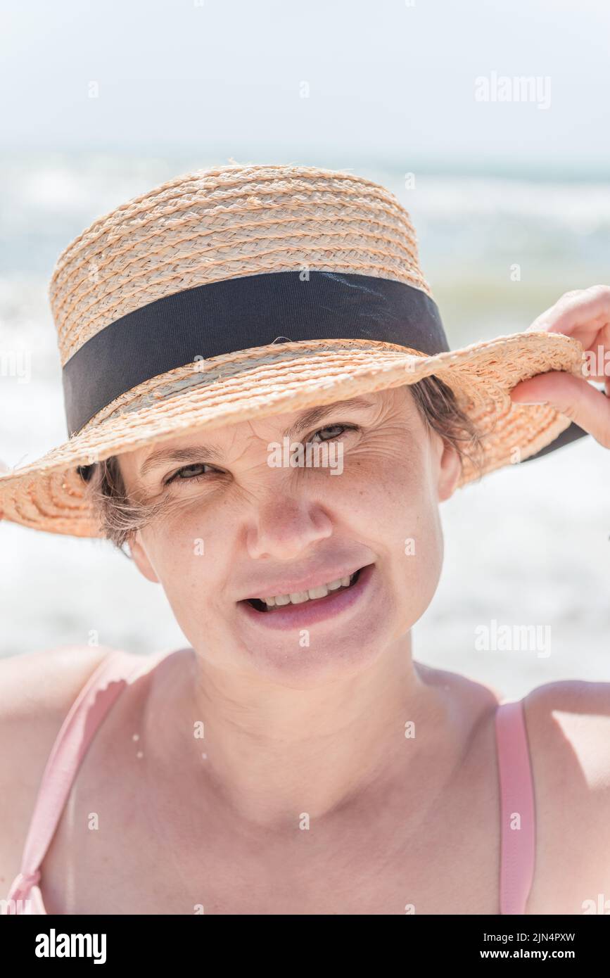 Glückliche Frau in einem Strohhut lächelt aufrichtig an der Kamera, vor verschwommenem Hintergrund des Meeres. Das Konzept des Glücks. Stockfoto