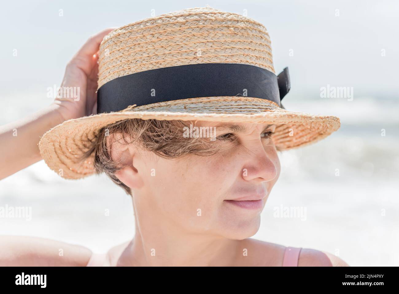 Frau in einem Strohhut auf dem verschwommenen Hintergrund des Meeres mit einem kleinen niedlichen Lächeln, das zur Seite schaut. Konzept der natürlichen Schönheit. Stockfoto