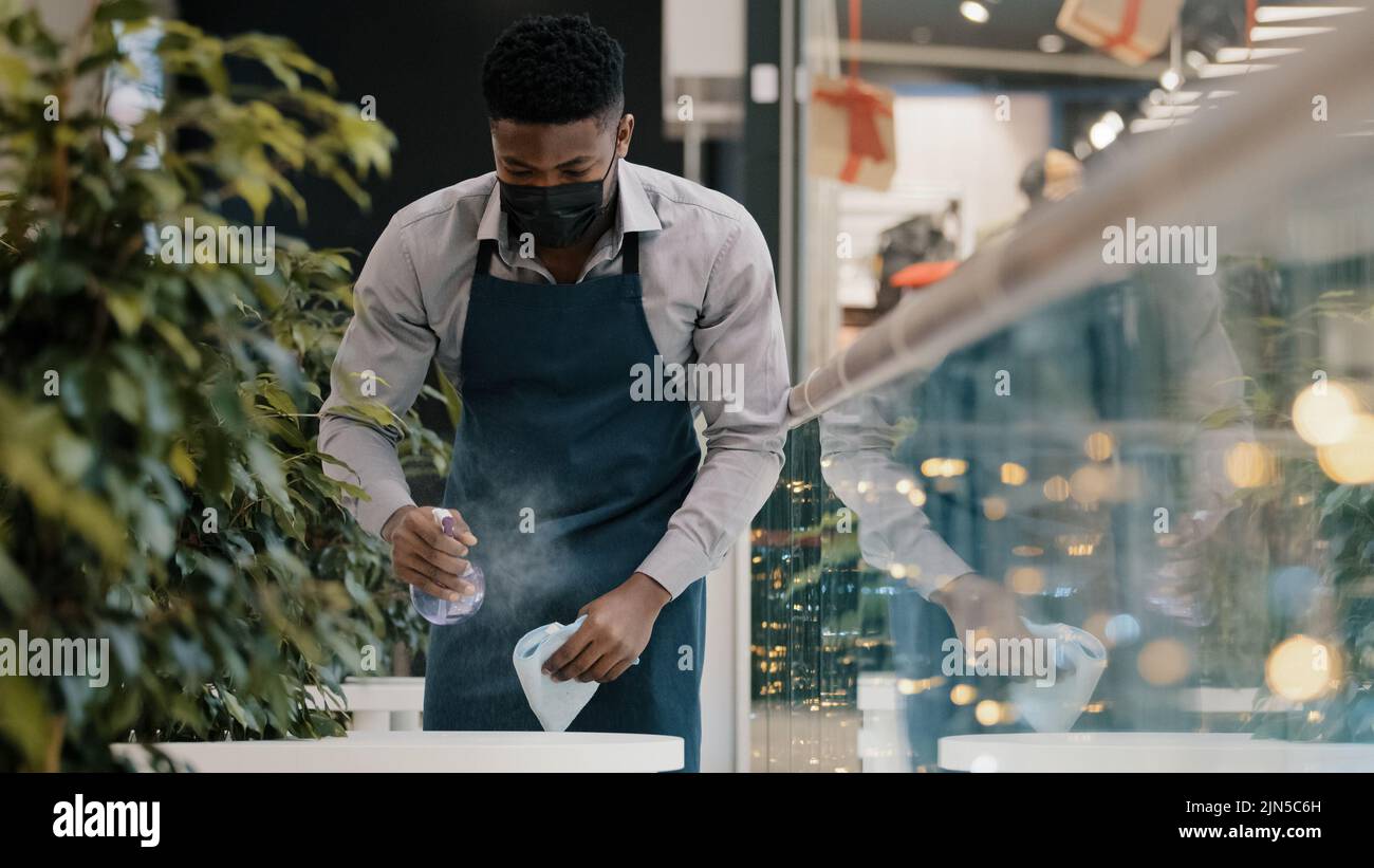 Junger männlicher Kellner in Schürze wischt Tisch Sprays Desinfektionsmittel Reiniger bereitet Café-Restaurant für die Eröffnung ernsthafte afroamerikanische Student in Medizin Stockfoto