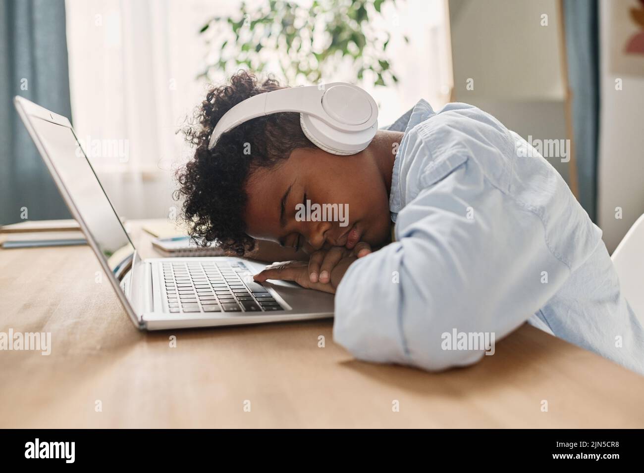 Afrikanischer müder Junge mit Kopfhörern, der nach dem Online-Unterricht zu Hause mit seinem Laptop an seinem Schreibtisch schlief Stockfoto