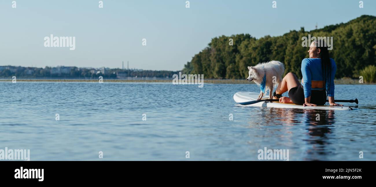 Junge Frau entspannt sich auf dem City Lake, während sie auf dem Sup Board mit ihrem Hund japanischer Spitz sitzt Stockfoto