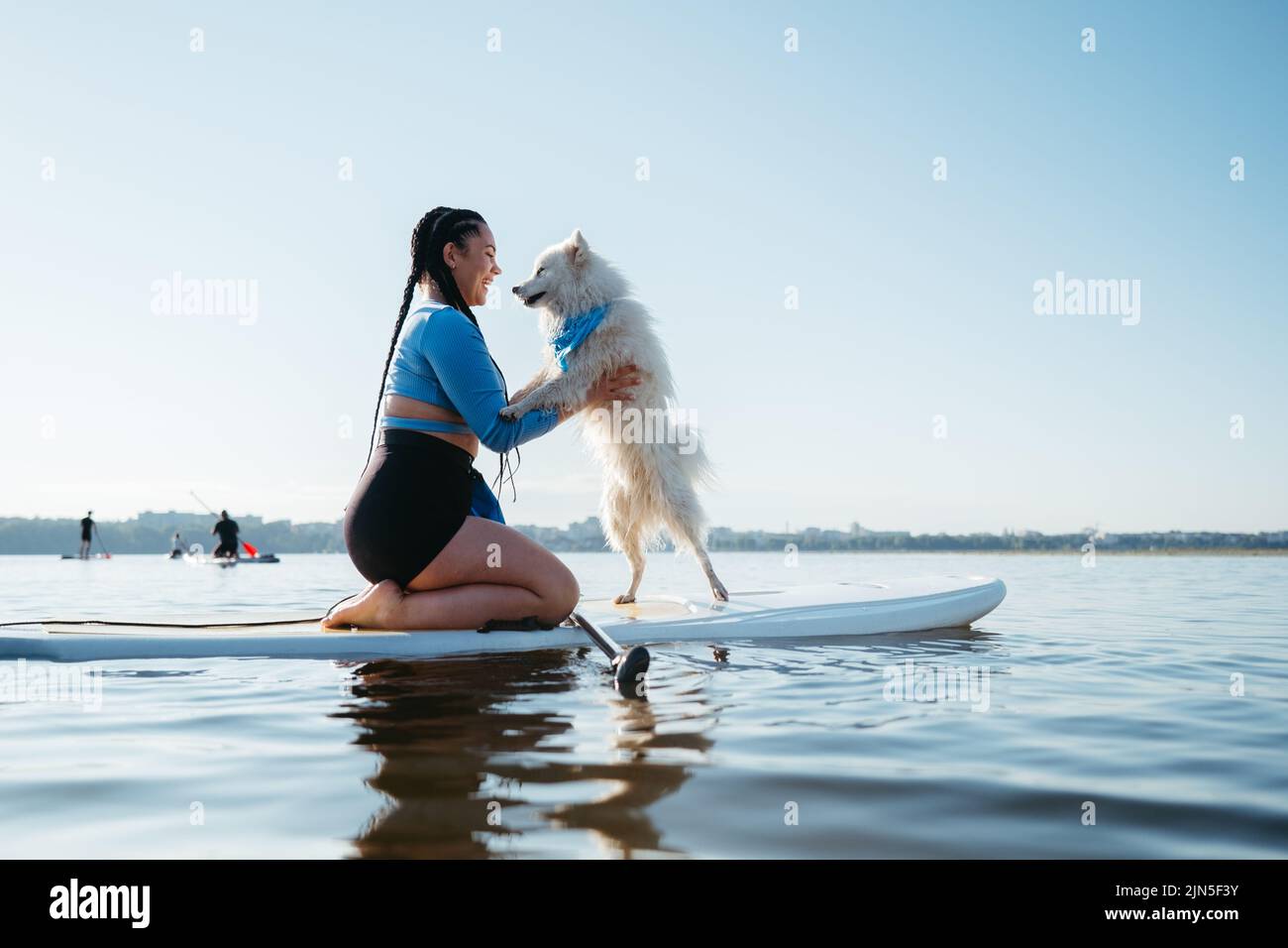 Glückliche junge Frau mit Locs umarmt mit ihrem Hund Japanischer Spitz, während sie auf dem Sup Board auf dem See sitzt Stockfoto