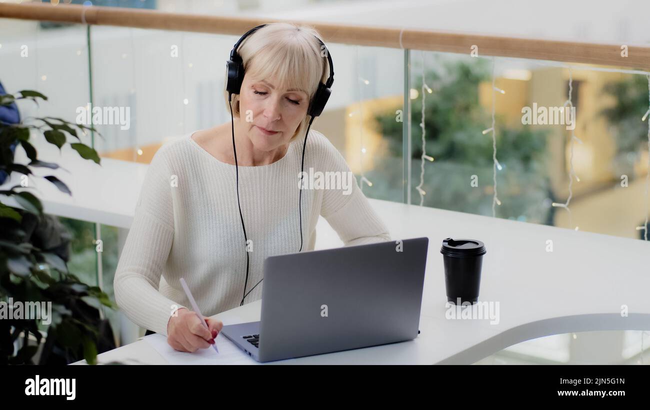 Reife kaukasische Frau Geschäftsfrau Lehrer in Kopfhörern schaut auf Laptop-Bildschirm nimmt Notizen schreibt Bericht Abstand lernen online Nachhilfe studieren Stockfoto