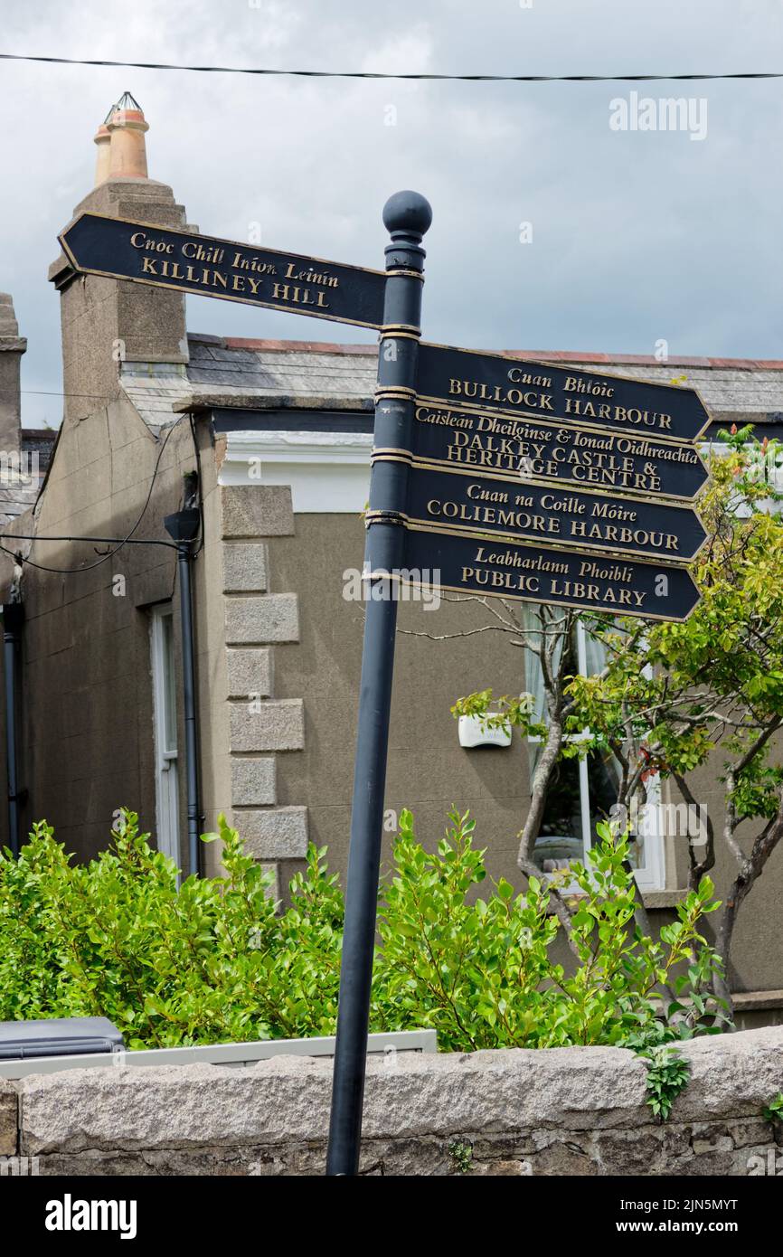 Vertikale Aufnahme eines Vintage-Schildes mit Anweisungen, die auf lokale Touristenattraktionen in Dalkey Stadt zeigen. Zweisprachiger Text in irischer und englischer Sprache. Stockfoto