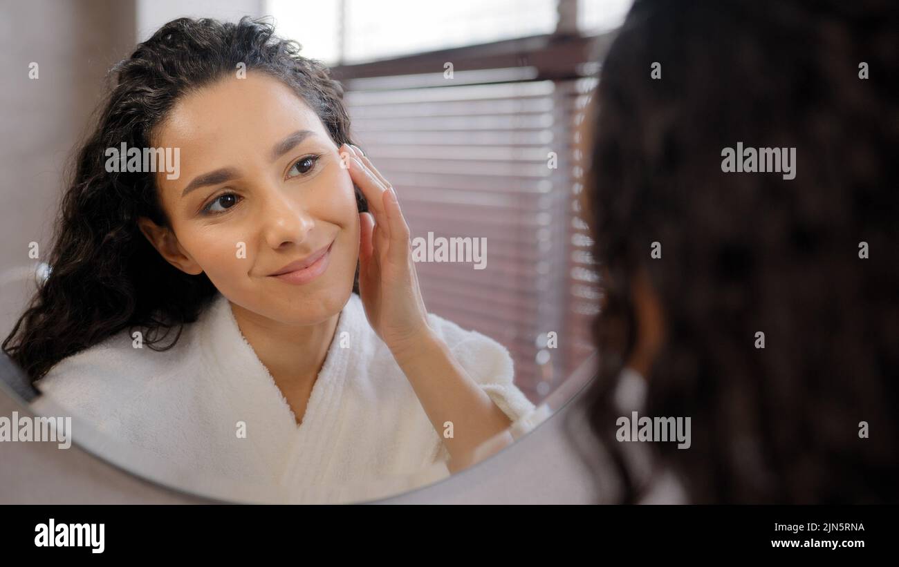 Junge attraktive Frau im Bademantel sieht im Spiegel im Badezimmer berührt mit Feuchtigkeit versorgt Gesicht nach kosmetischen Verfahren zu Hause genießen gesund sauber weich Stockfoto