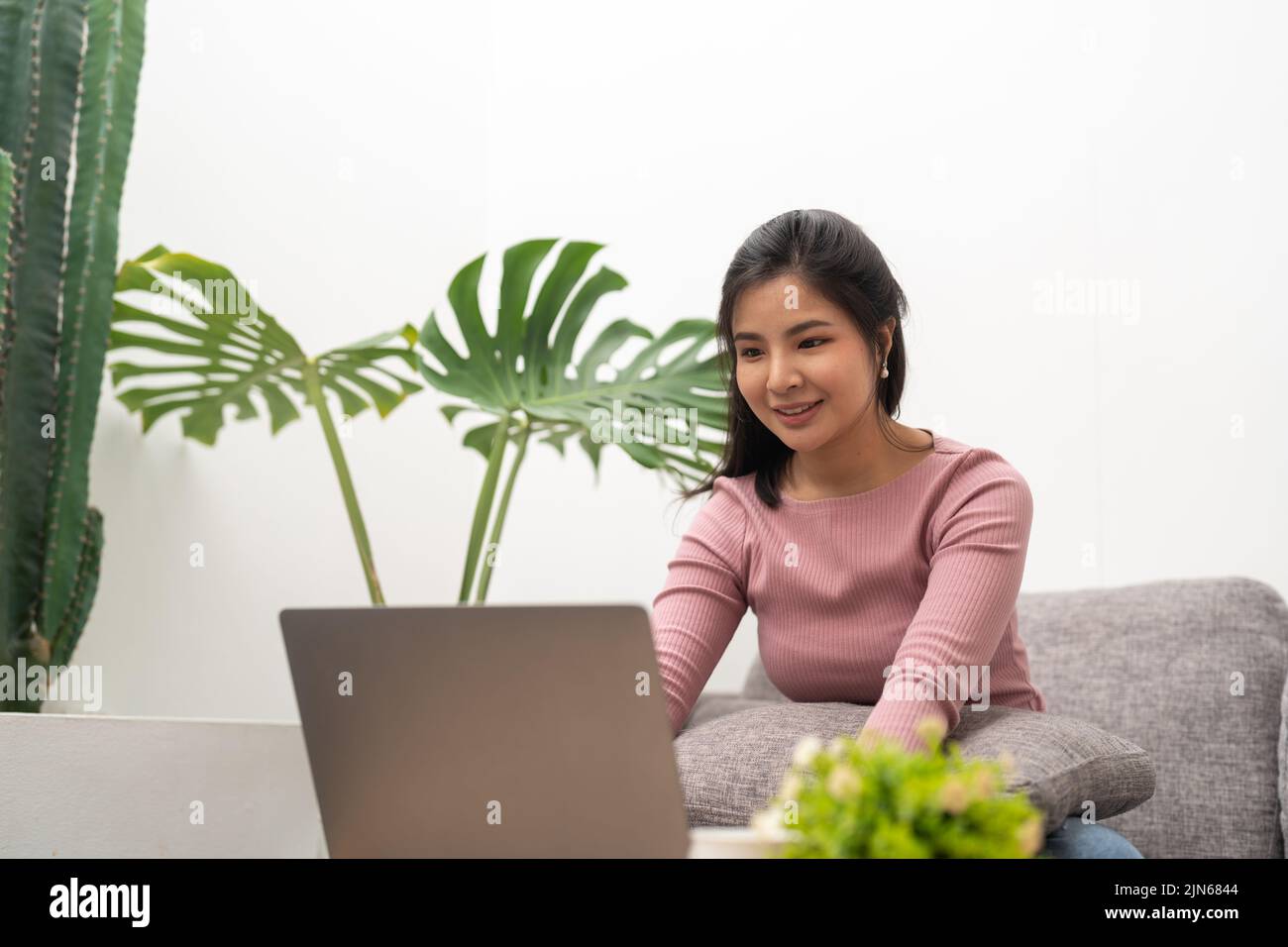 Fröhliche asiatische Frau mit Laptop, während sie auf dem Sofa im Wohnzimmer sitzt Stockfoto