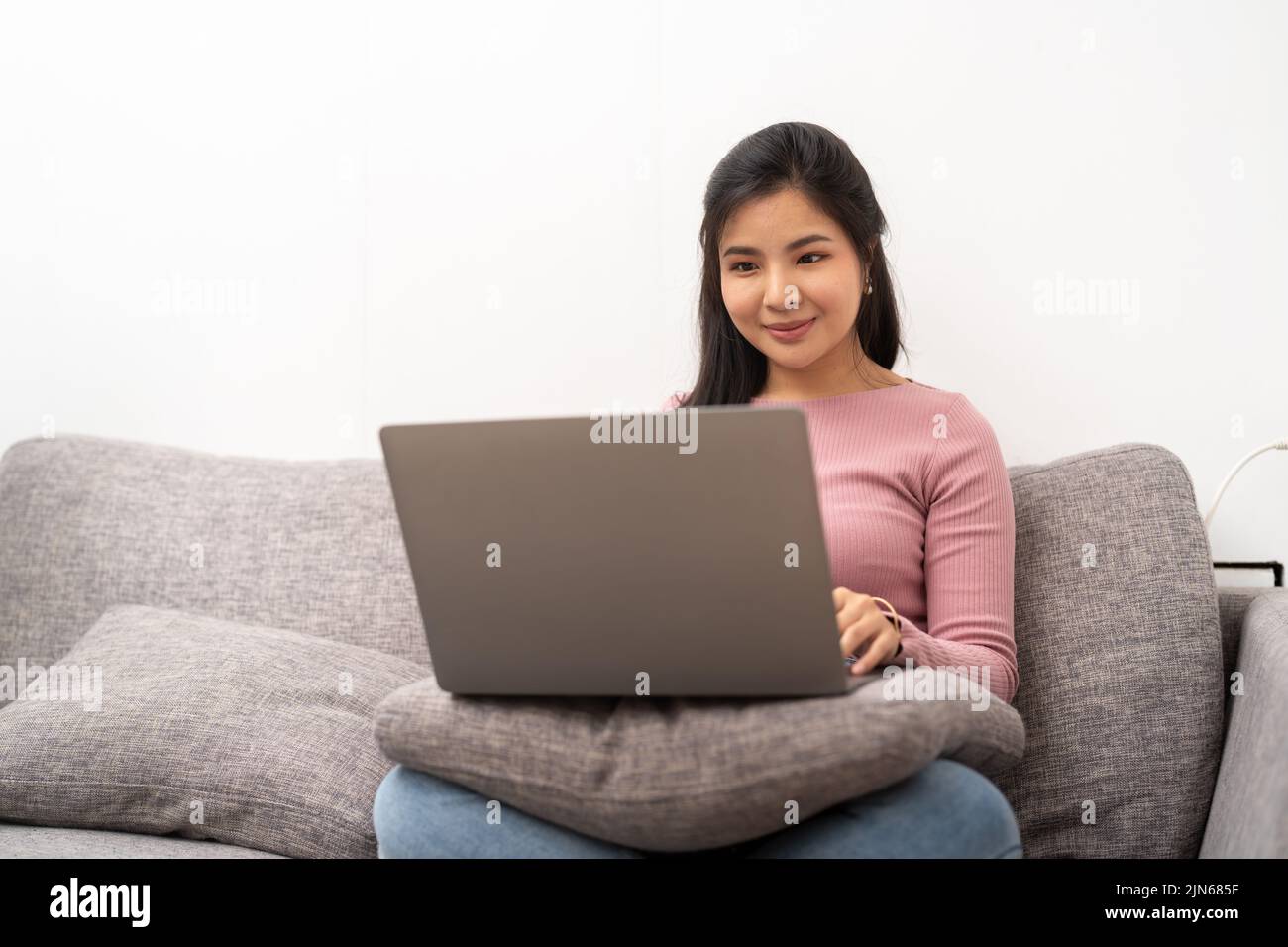 Fröhliche asiatische Frau mit Laptop, während sie auf dem Sofa im Wohnzimmer sitzt Stockfoto