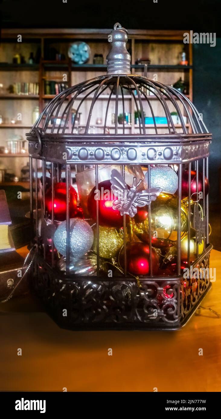 Draht- und Metallkäfig voller Weihnachtsschmuck und Lichter vor einem dunklen, verschwommenen Raumhintergrund mit Regalen und neben Büchern auf einem Tisch Stockfoto