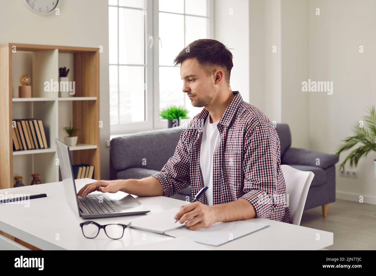 Konzentrierter junger Mann, der im Heimbüro ferngesteuert am Laptop arbeitet und Notizen im Notebook aufnimmt. Stockfoto