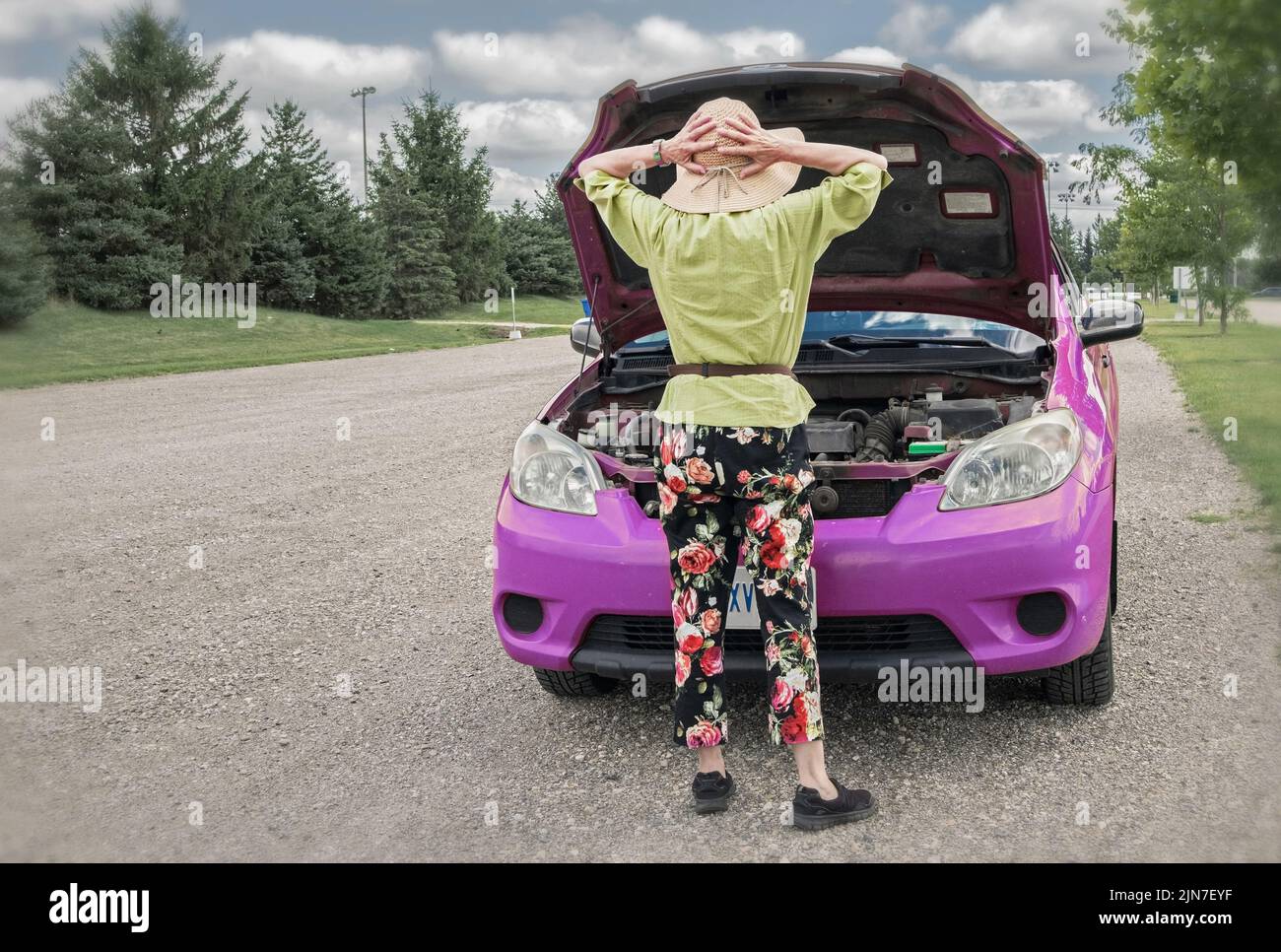 Von hinten, eine Frau, die wahrscheinlich verzweifelt sehen will, wie ihr Auto mitten im Nirgendwo zusammenbricht. Sie trägt einen breitkrempigen Strohhut und eine blumige Hose. Stockfoto