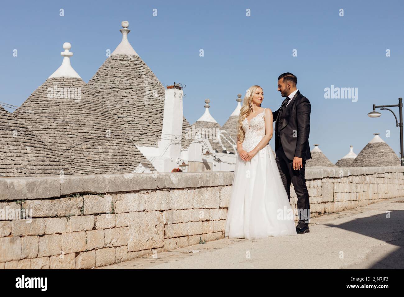 Glückliches Hochzeitspaar, das auf der Straße in der Nähe der konischen Dächer von Trullo steht und Augen trifft. Junge Braut umarmt sich mit Bräutigam. Stockfoto