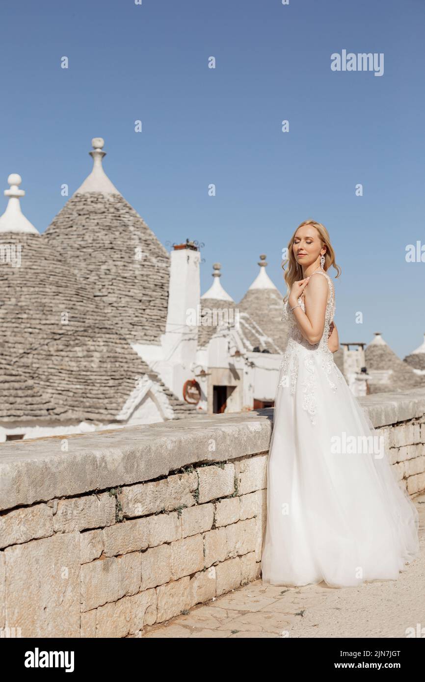 Seitenansicht der jungen Frau Braut mit langen, hellen Haaren in weißem Hochzeitskleid, stehend in der Nähe von konischen Dächern von Trullo. Stockfoto