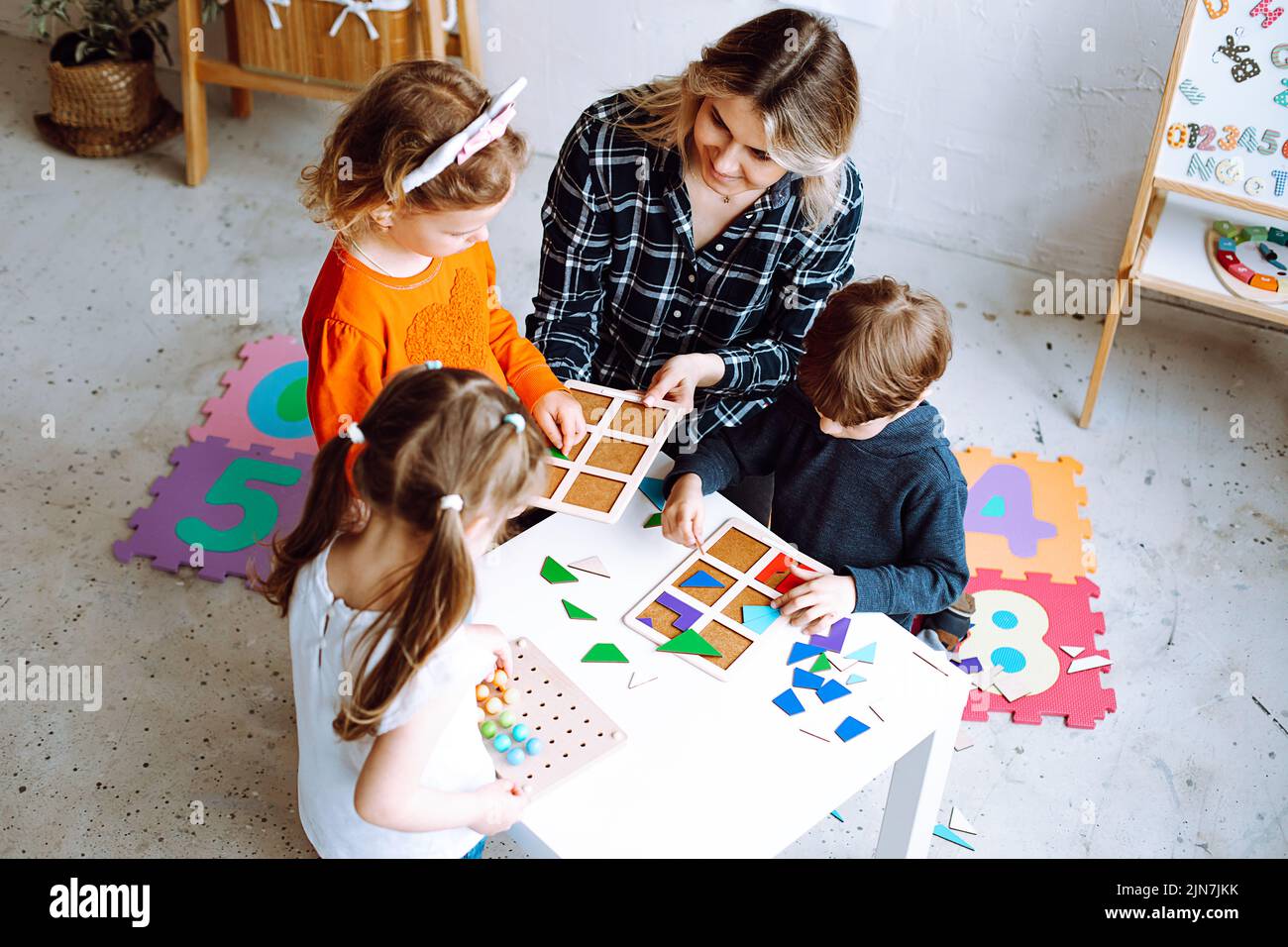 Draufsicht auf die Gruppe der Vorschulkinder. Junge blonde Frau hilft Schülern, Brettspiele zu spielen und geometrische Figuren zu sammeln. Stockfoto