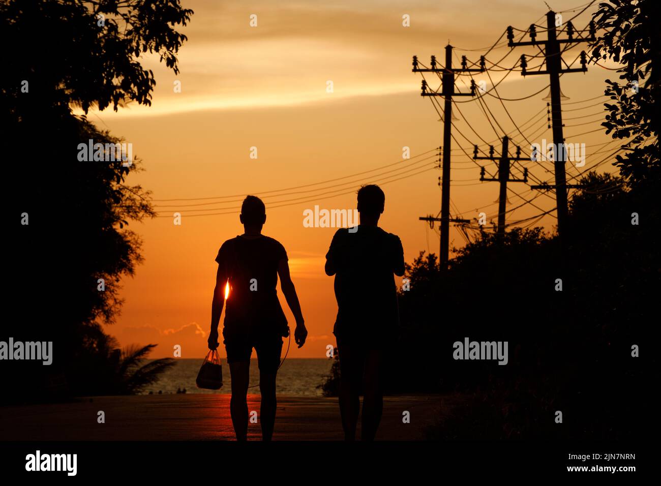 Eine Frau und ein Mann in Silhouette gehen entlang einer thailändischen Inselstraße zu einem wunderschönen tropischen Strand und Sonnenuntergang am Meer Stockfoto