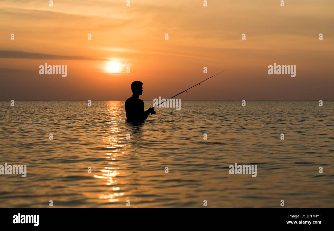 Ein eingeflügelter Fischer ist im orangefarbenen Glanz eines tropischen Sonnenuntergangs silhouettiert, als er im Wasser steht und seine Angelköder abwirft Stockfoto
