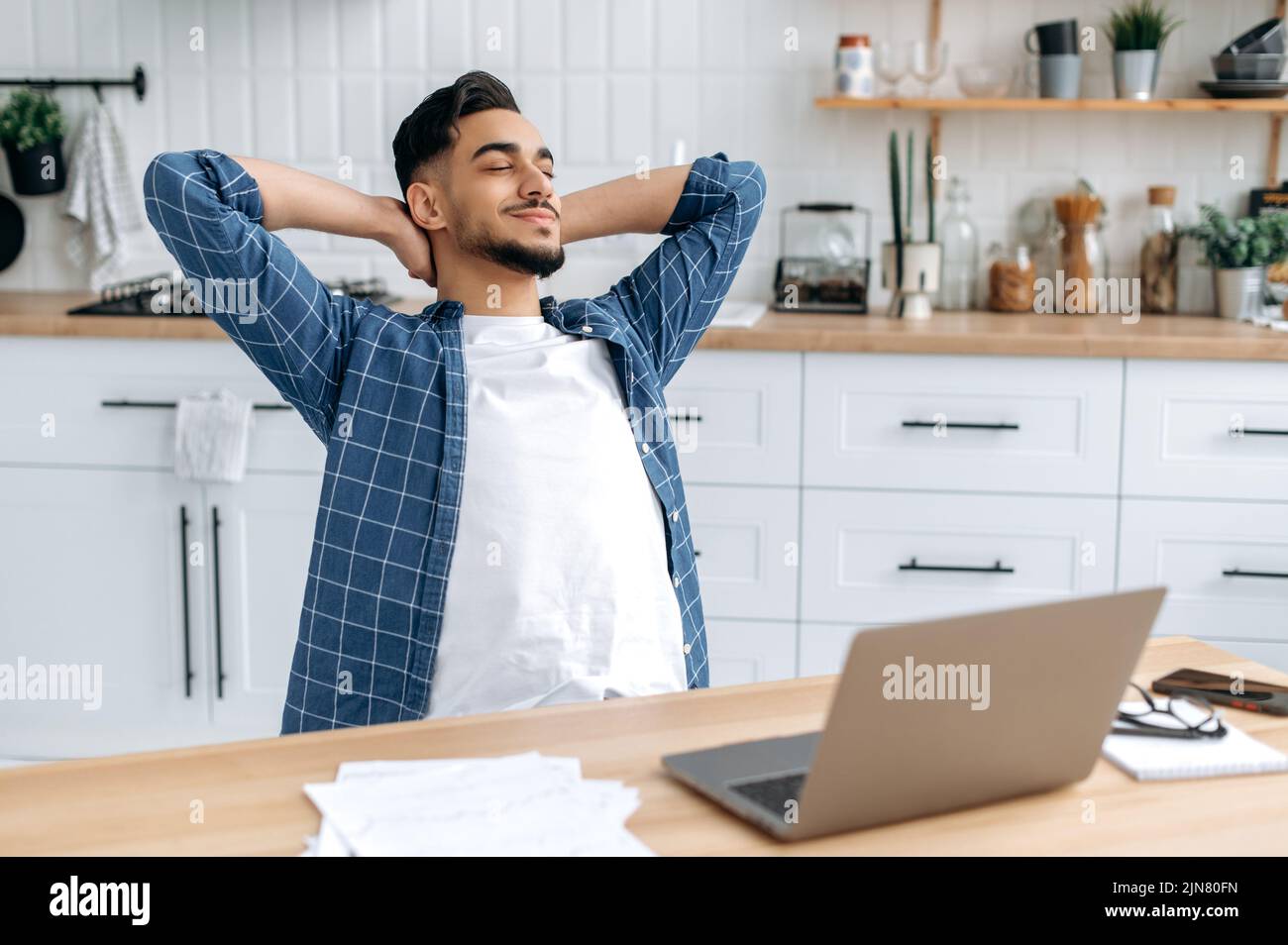 Entspannter, ruhiger araber oder inder, Freiberufler, arbeitet von zu Hause aus, sitzt in der Küche am Arbeitsplatz, macht eine Pause von der Arbeit, legt seine Hände hinter den Kopf, schließt seine Augen, träumt von Ruhe, lächelt Stockfoto