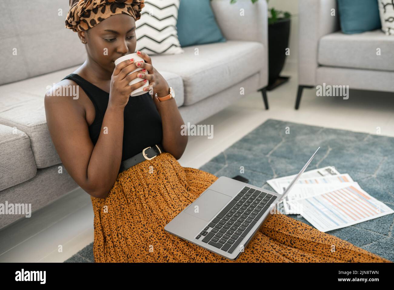 Wunderschöne afrikanische Frau, die in der Lounge auf dem Boden sitzt, zu Hause auf dem Laptop sitzt und Kaffee in der Hand trägt, einen traditionellen Kopftuch trägt und an einem Coff nippt Stockfoto