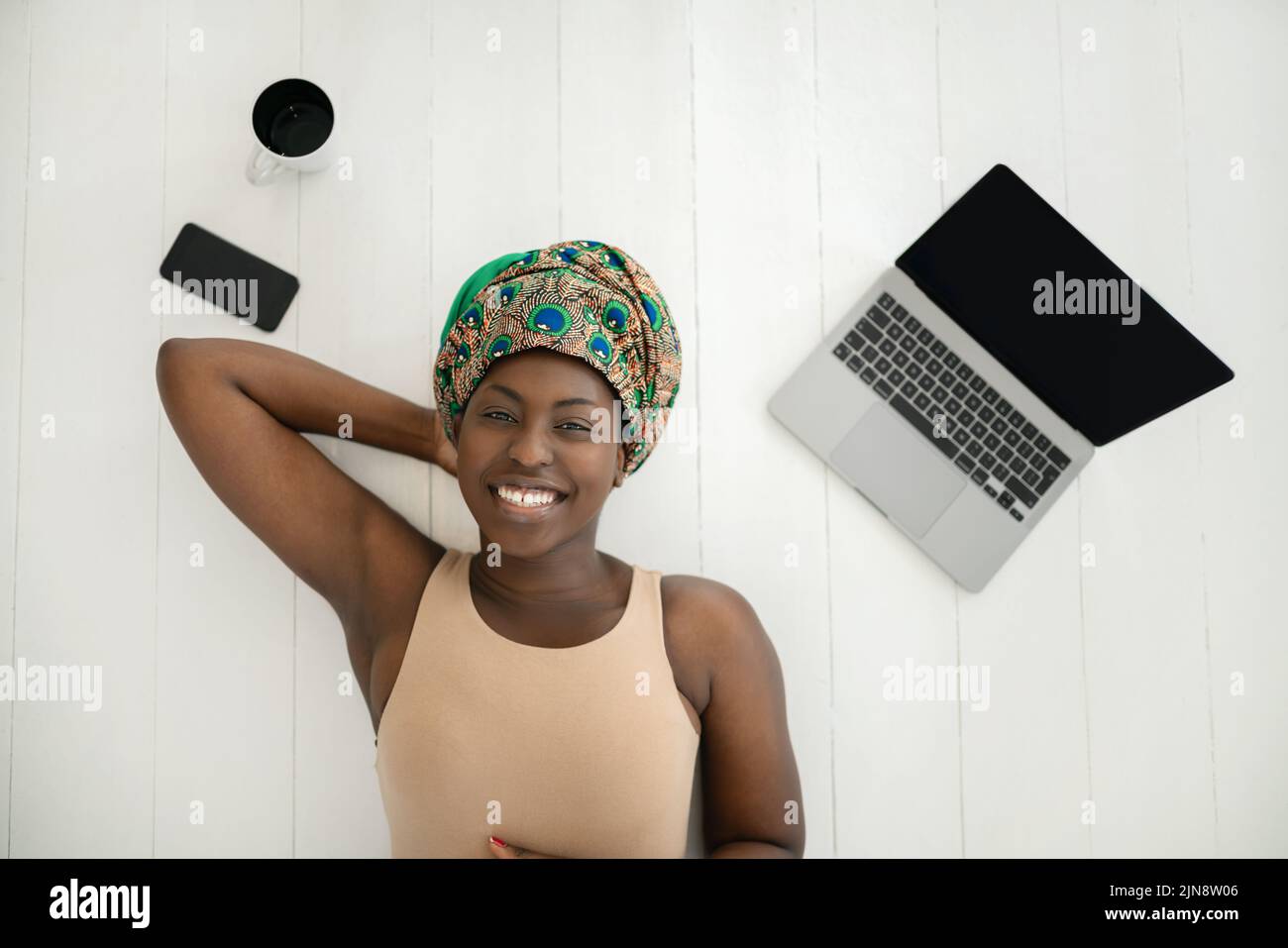 Draufsicht der schönen jungen afrikanischen Frau, die auf weißem Holzboden liegt und einen traditionellen Schal mit Kopfband trägt. .Mit Laptop, Handy und Tasse o Stockfoto