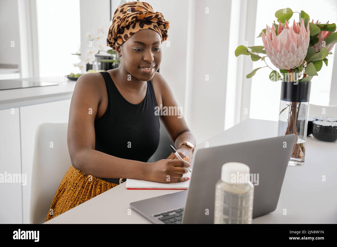 Afrikanische Frau, die von zu Hause aus studiert, auf einen Laptop schaut und Notizen schreibt. Laptop und Smartphone auf dem Schreibtisch des Heimbüros Stockfoto