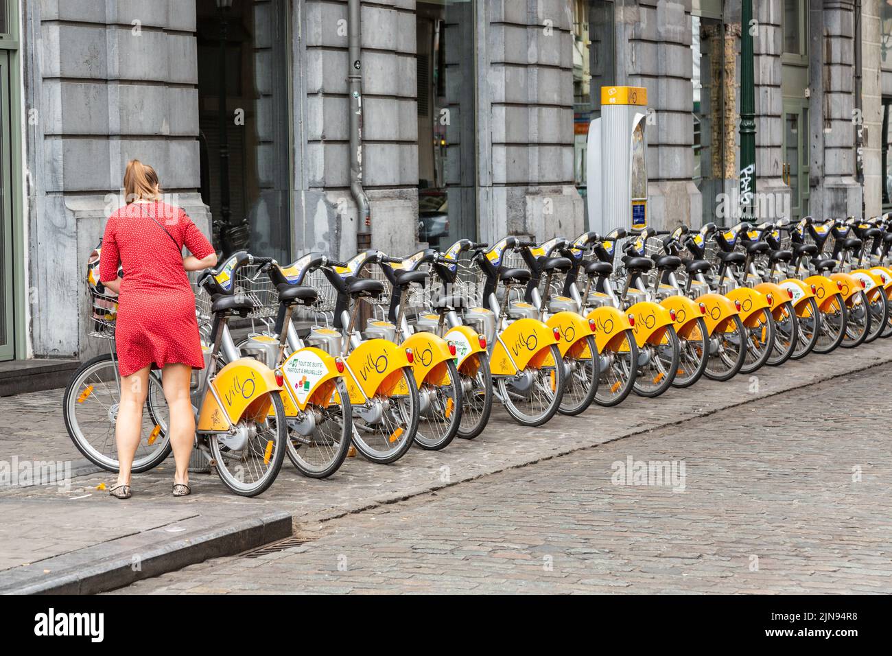 Dame in einem roten Kleid, die in einer Straße in Brüssel ein gemeinsames Fahrrad in Besitz nimmt Stockfoto