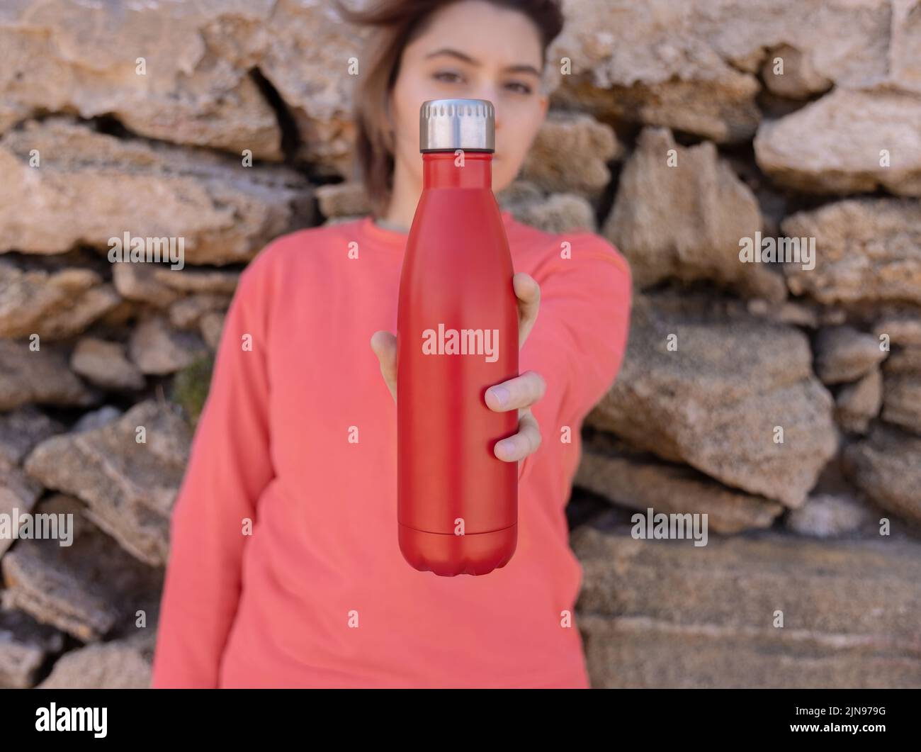Ernsthafter Teenager im pinken Hoodie mit roter Wasserflasche in der Hand, die straight aussieht. Kurzhaarige Teenager-Mädchen, die sich im Freien in der Nähe einer Steinmauer aufhalten. Kleidung und r Stockfoto