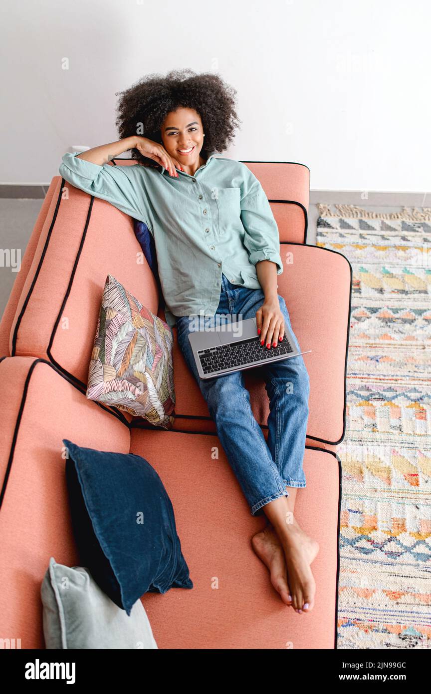 Draufsicht auf dunkle Haut lockige junge Frau im Internet surfen oder SMS mit Freunden online, mit Laptop sitzend auf dem gemütlichen Sofa, arbeitet freiberufliche Mitarbeiterin aus der Ferne, schaut auf die Kamera und lächelt Stockfoto