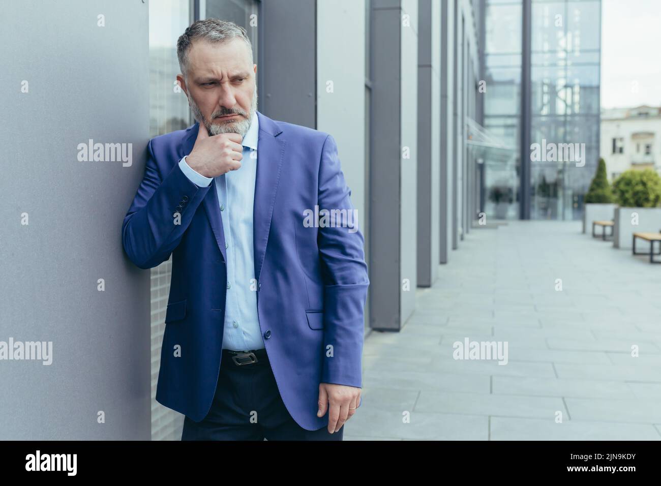 Leitender grauhaariger Mann vor dem Bürogebäude, stehend und denkend, verwirrt und traurig Geschäftsmann Chef in Business Anzug, über Entscheidung Stockfoto