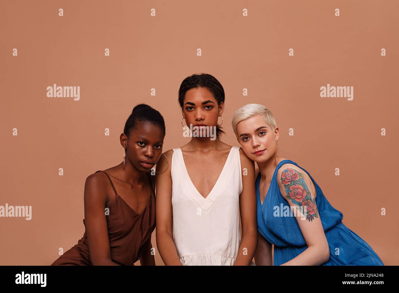 Drei schöne Frauen mit unterschiedlichen Hautfarben betrachten die Kamera. Junge Frauen in Kleidern sitzen auf braunem Hintergrund. Stockfoto