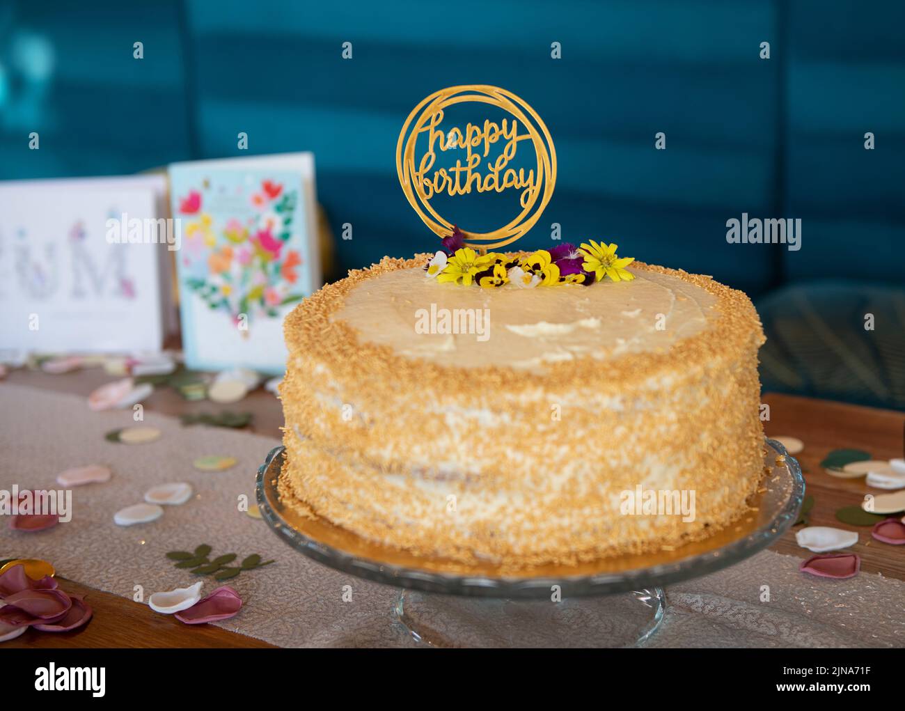 Geburtstagstorte mit Buttercreme-Verzierung und Streuseln auf einem Tisch mit Geburtstagskarten und Blütenblatt-Dekorationen Stockfoto