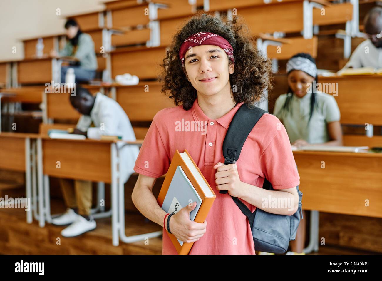 Porträt eines Studenten mit Rucksack und Buch Blick auf die Kamera, während im Auditorium der Universität mit Klassenkameraden im Hintergrund stehen Stockfoto