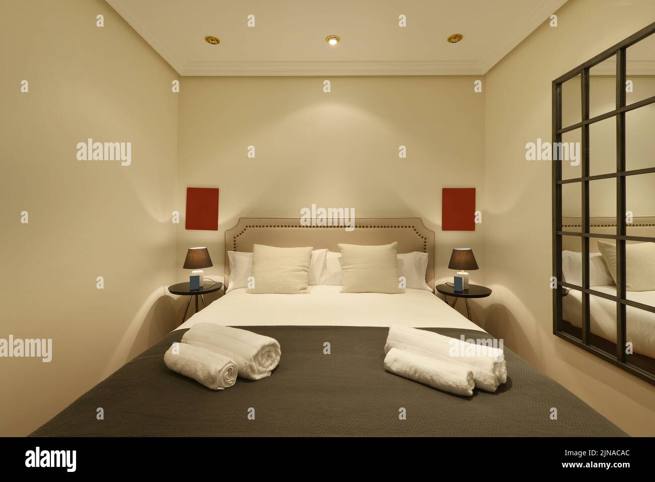 Schlafzimmer mit Kopfteil, gepolstert mit hellem Stoff, Kissen im gleichen Stil, Beistelltischen und zwei Lampen sowie einem Spiegel an einer Wand Stockfoto