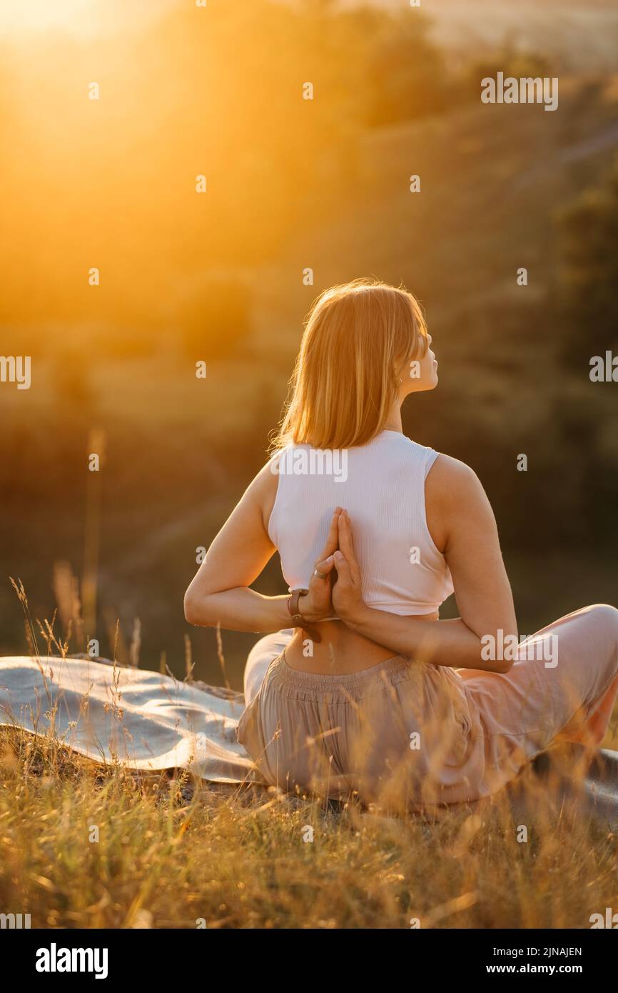Junge Frau mit gefalteten Händen auf dem Rücken meditiert und genießt den wunderschönen Sonnenuntergang im Freien Stockfoto