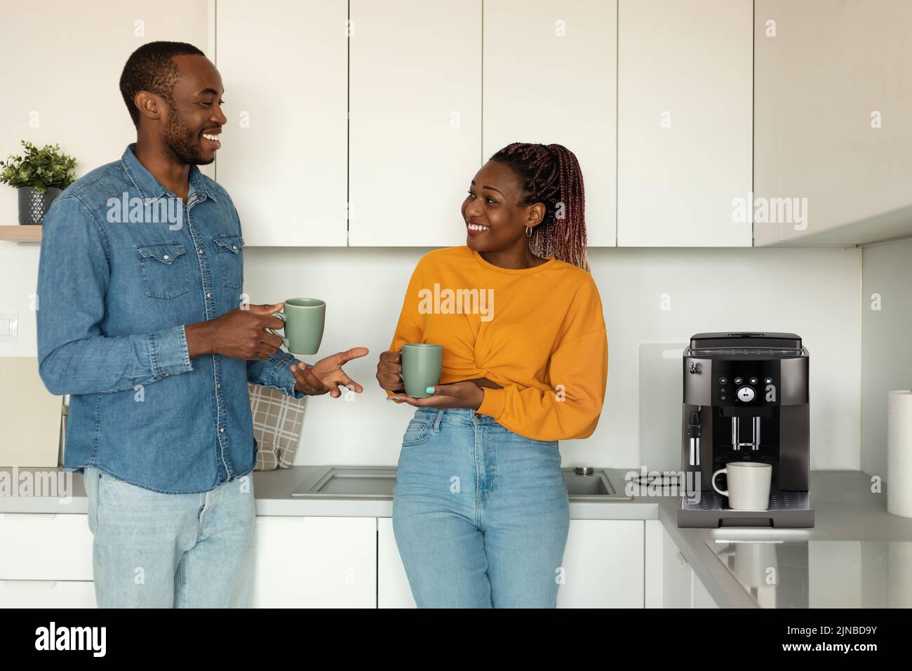 Junge afroamerikanische Ehegatten trinken Morgenkaffee und reden, während sie in der Nähe einer Kaffeemaschine in der Küche stehen Stockfoto