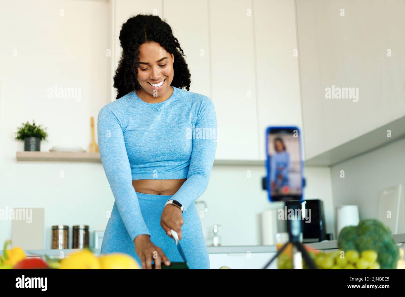 Glückliche afroamerikanische Dame in Sportswear, die Video auf der Smartphone-Kamera aufzeichnete und frischen Salat in der Küche zubereitete Stockfoto