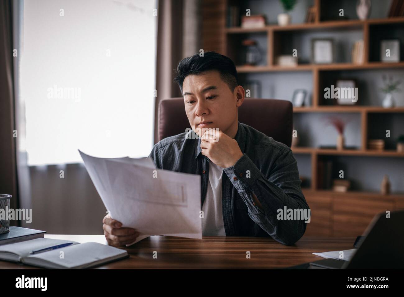 Konzentrierter, ruhiger Chinese mittleren Alters am Tisch arbeitet mit Dokumenten im Innenleben des Heimbüros Stockfoto