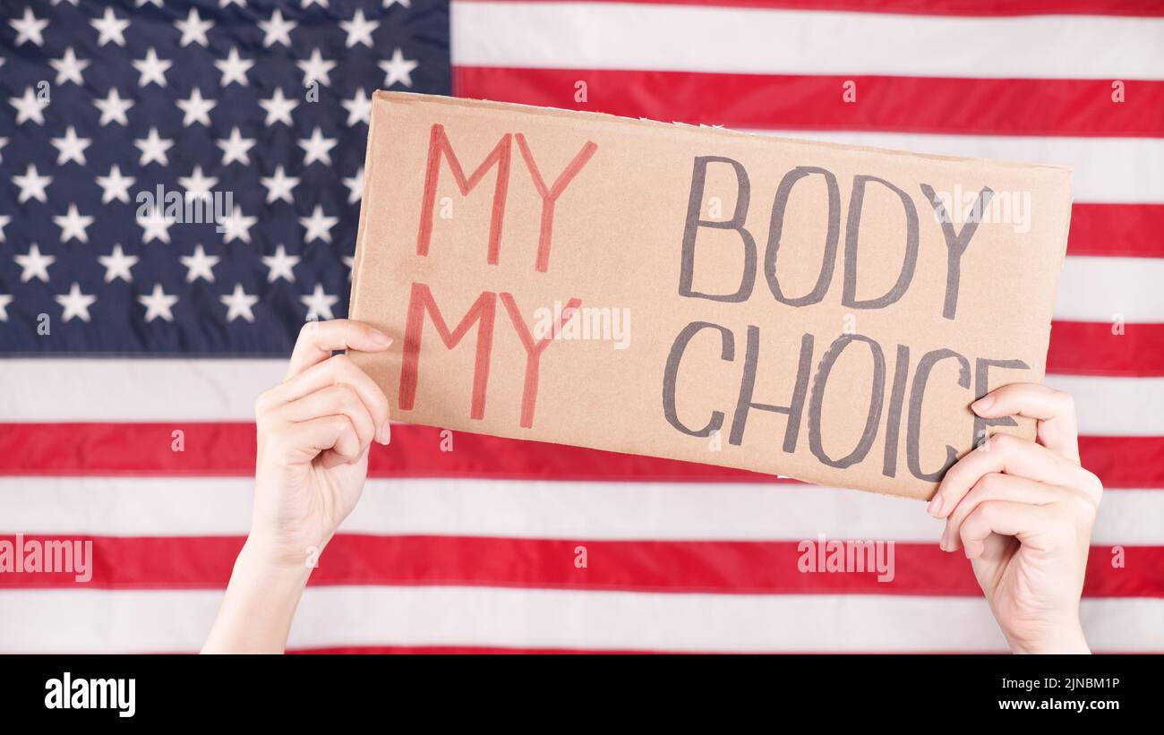Junge Frau Protesterin hält Karton mit My Body My Choice Zeichen gegen USA Flagge auf Hintergrund. Mädchen protestiert gegen Anti-Abtreibungsgesetze. Feministisch Stockfoto