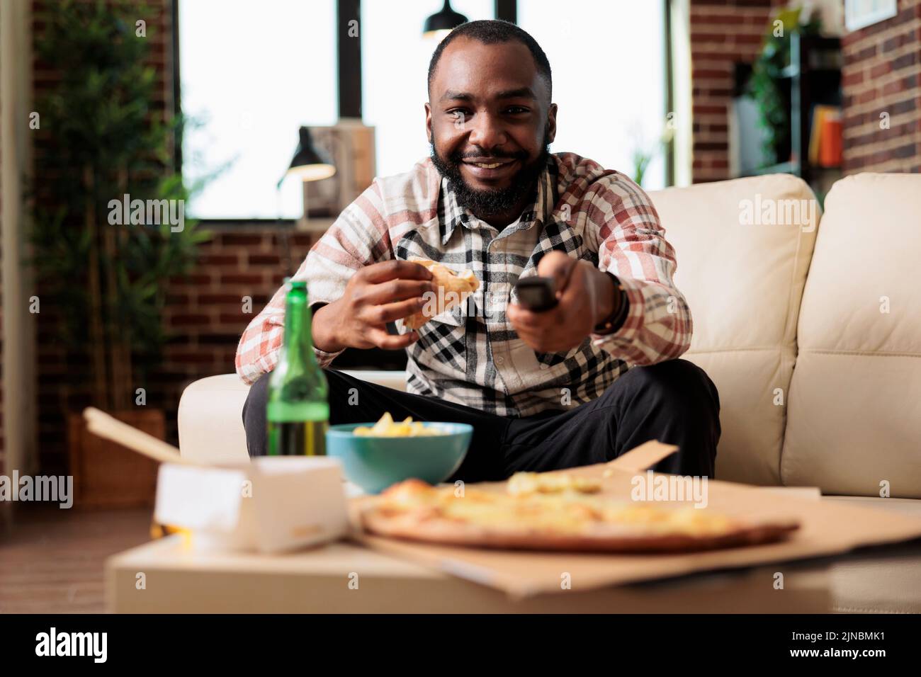 afroamerikanischer Mann, der die fernbedienung des fernsehers benutzte, um den Sender zu wechseln und ein Stück Pizza von der Lieferung mit Schnellimbiss zu essen. Spaß mit Essen zum Mitnehmen und Bier, während er sich einen Film zusieht. Stockfoto