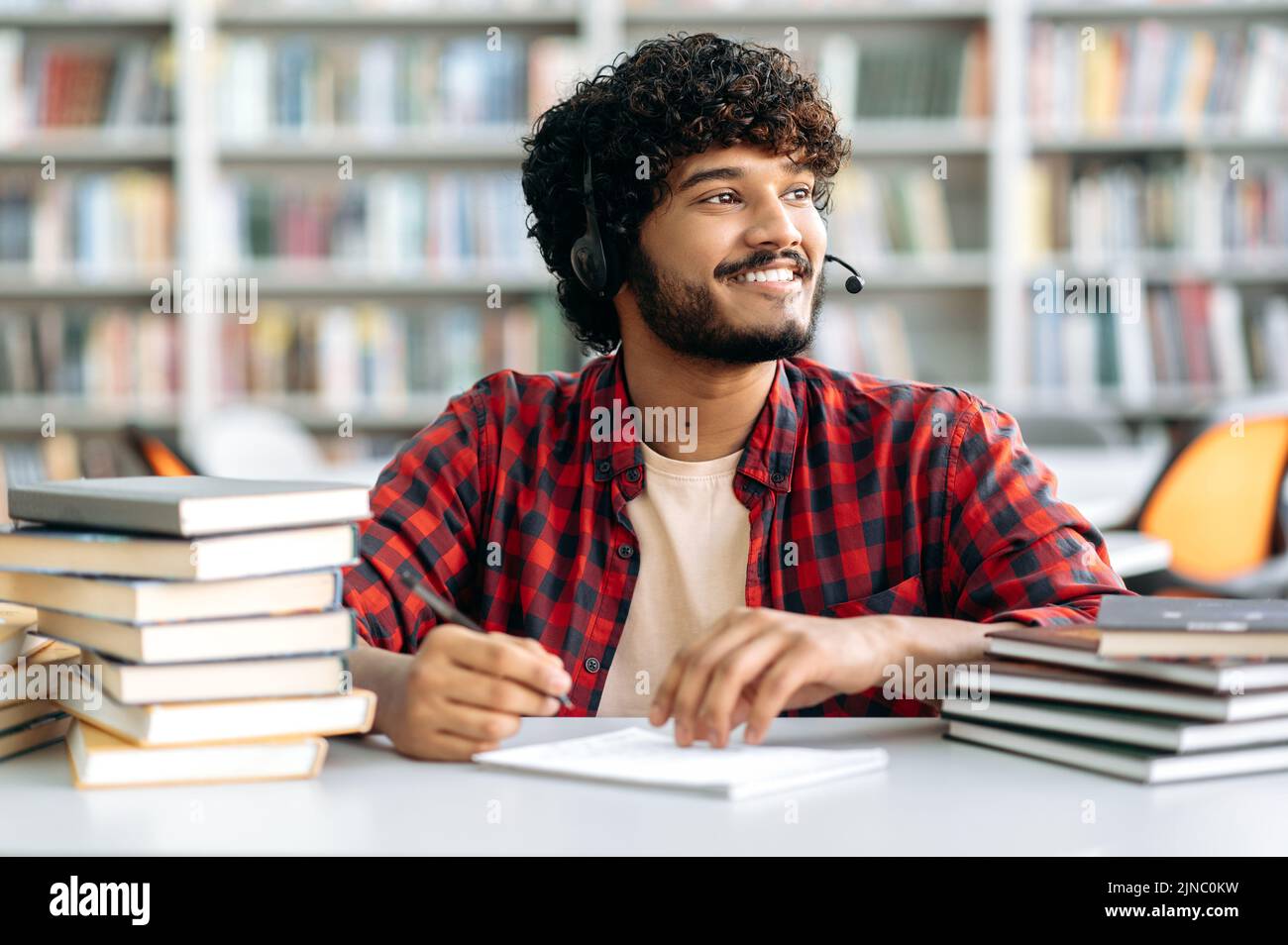 Positiver arabischer oder indischer Student mit lockigen Haaren, mit einem Headset, sitzt mit Büchern an einem Tisch in der Bibliothek, hört sich ein Online-Webinar an, hält Vorträge, schaut weg, träumt, denkt, Lächelnd Stockfoto