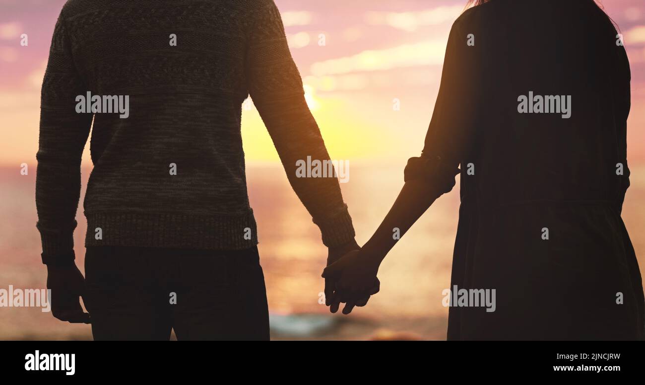 Wahre Liebe bei Sonnenuntergang. Rückansicht eines unerkennbaren, liebevollen Paares, das sich bei Sonnenuntergang am Strand die Hände hält. Stockfoto