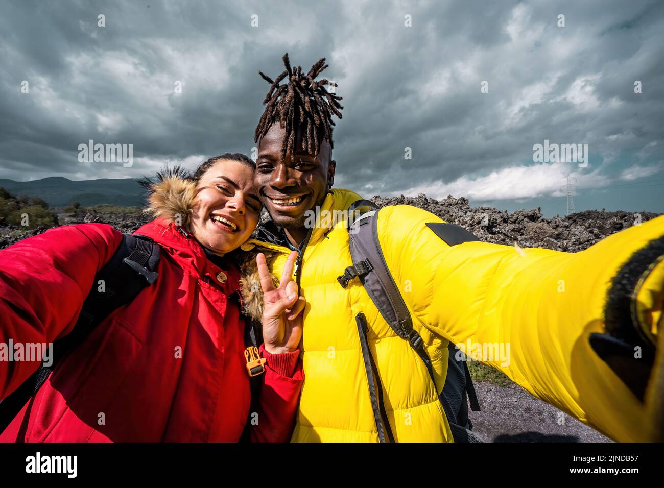 Multiethnisches Paar junger Wanderer, die Selfie-Erinnerungen auf einer wolkigen und vulkanischen Lavasteinlandschaft machen - afrikanischer Freund mit kaukasischer Freundin Stockfoto