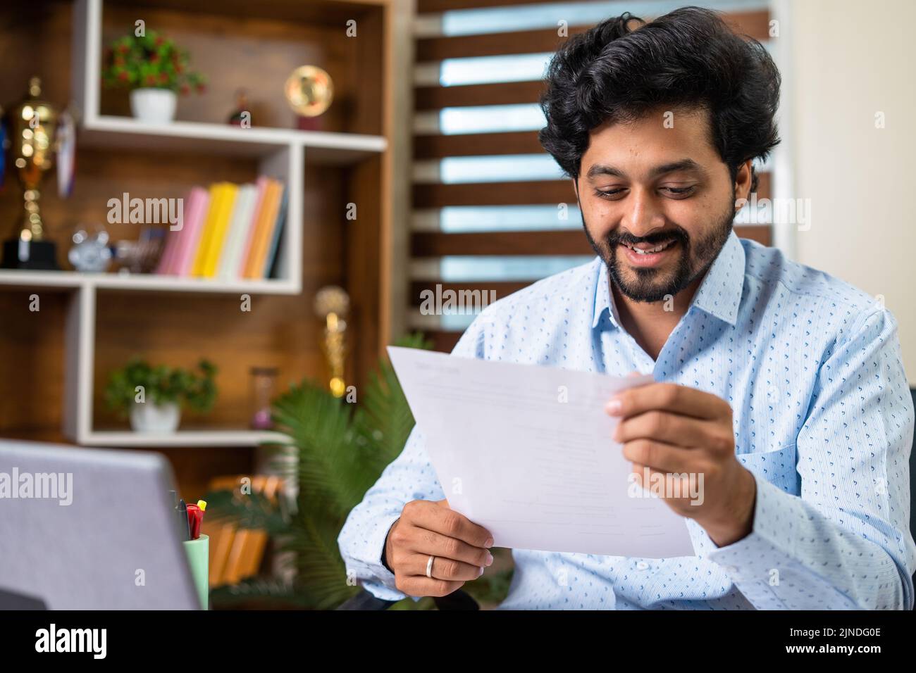 Glücklich lächelnder junger Mann, der Angebot oder Werbebrief liest, während er im Büro sitzt - Konzept der Leistung, Kreditgenehmigung und Arbeitsförderung Stockfoto