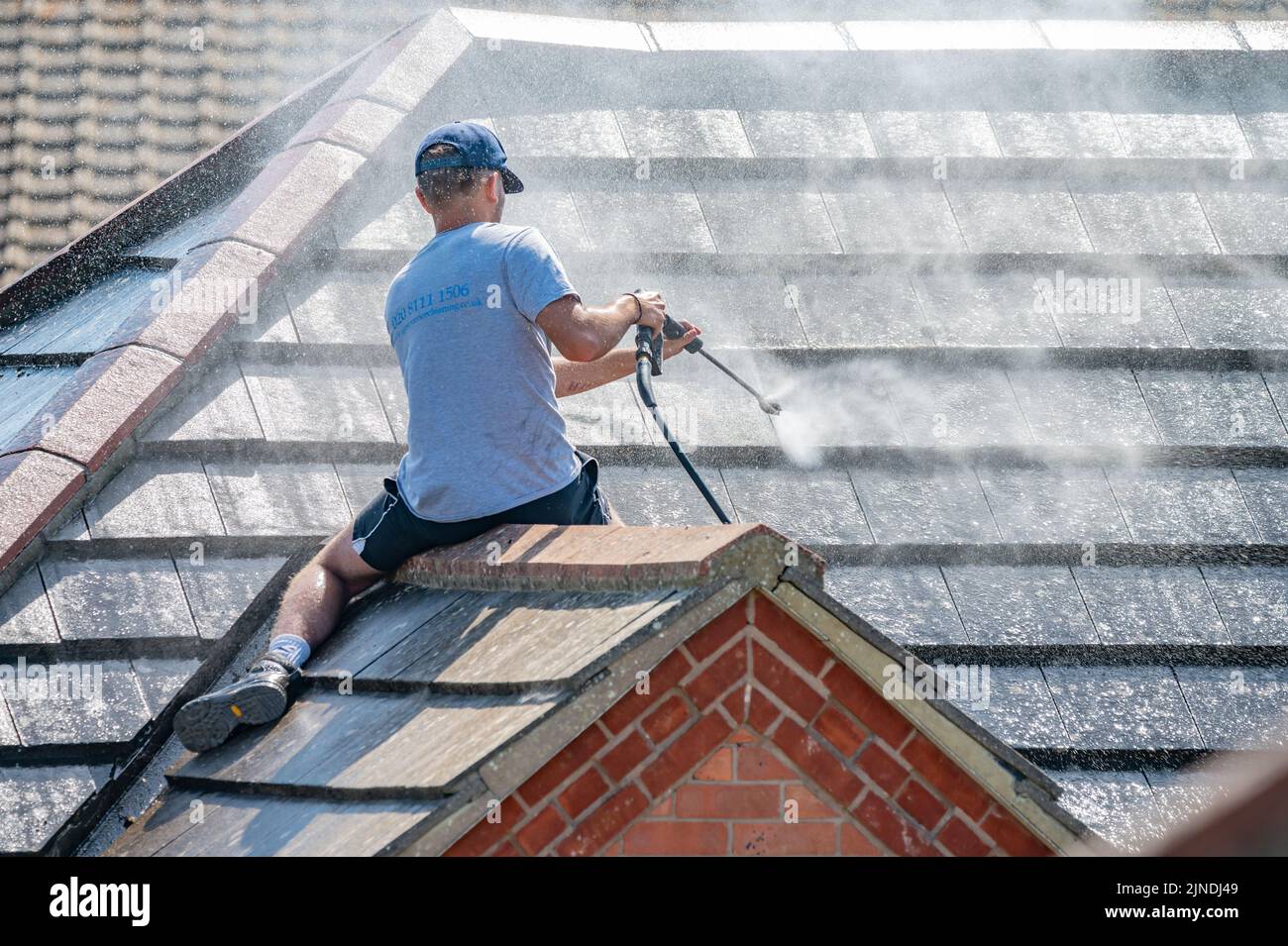 Mann sitzt auf einem Ziegeldach eines Hauses, benutzt einen Hochdruckreiniger an einem Schlauch, feuert einen Wasserstrahl ab, um das Dach und die Fliesen zu reinigen. Stockfoto