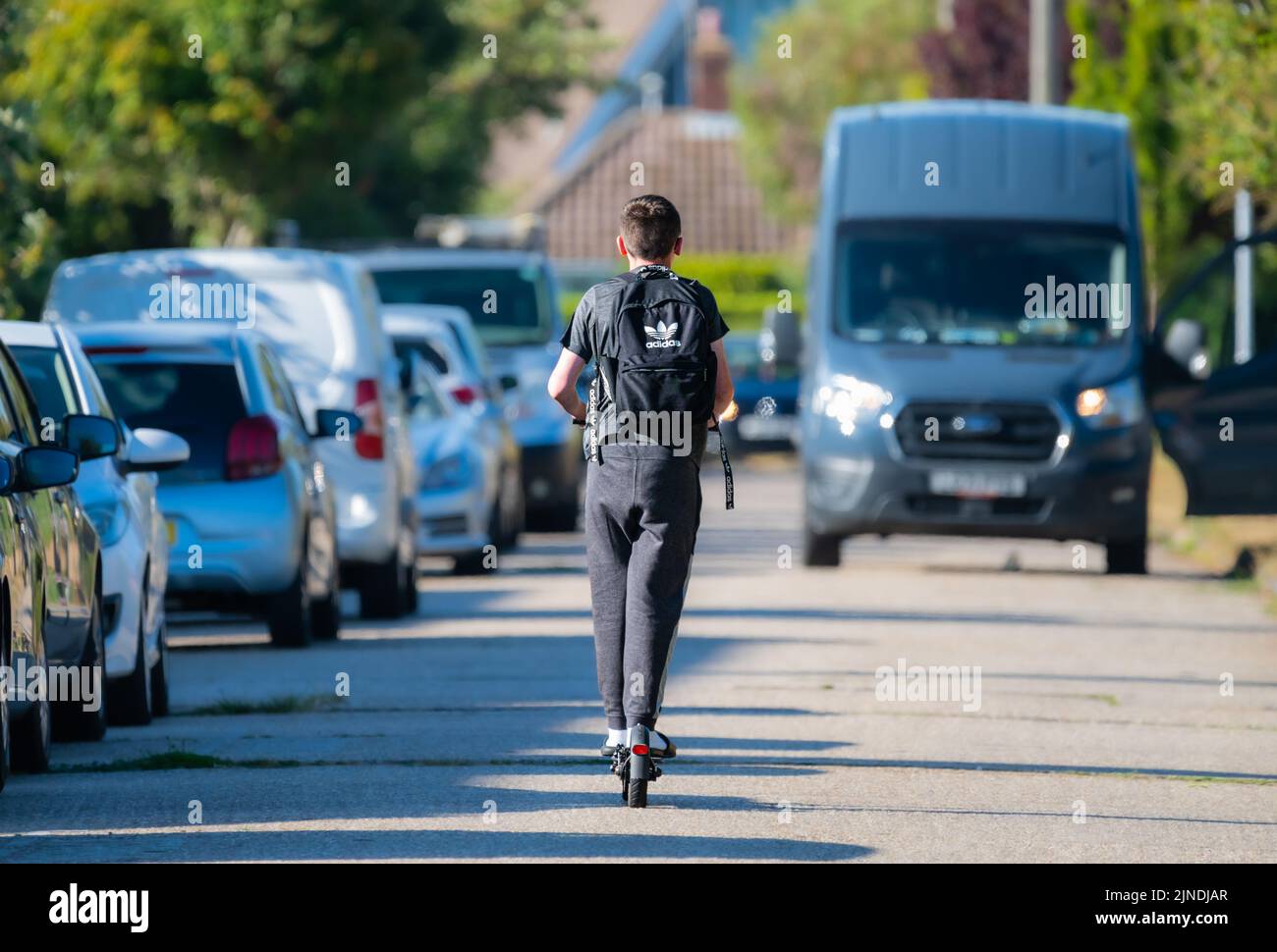 Ein Junge, der in England auf einer britischen Straße mit geparkten Autos auf einem Elektroroller oder E-Scooter fährt. Stockfoto