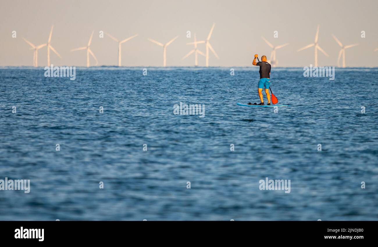 Ein Mann steht auf dem Paddleboard und paddelt an einem heißen Tag im Sommer in England, Großbritannien, mit Windturbinen von einer Offshore-Windfarm im Hintergrund. Stockfoto