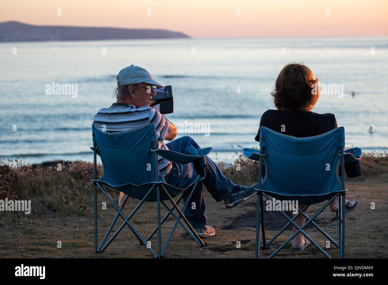 Zwei Personen sitzen auf Campingstühlen, um den Sonnenuntergang in Godrevy, Cornwall, Großbritannien, zu beobachten und zu fotografieren Stockfoto