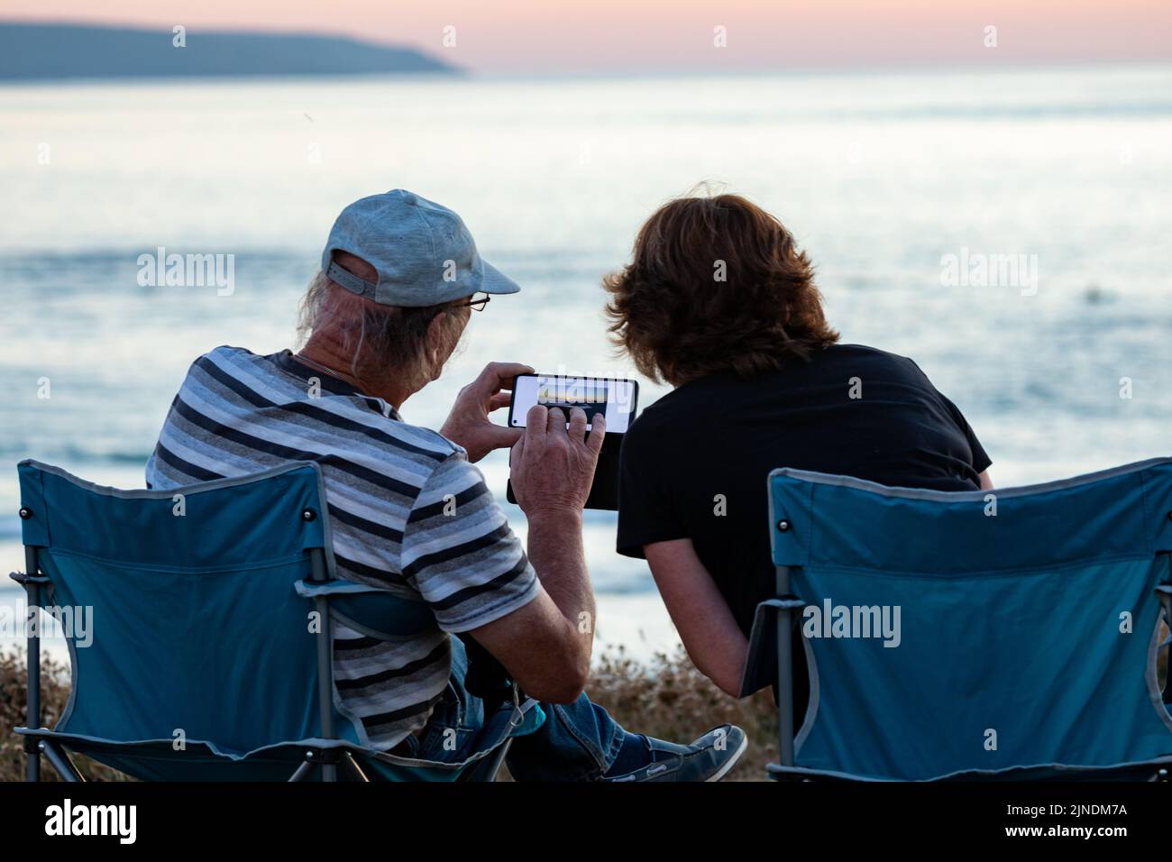 Zwei Personen sitzen auf Campingstühlen, um den Sonnenuntergang in Godrevy, Cornwall, Großbritannien, zu beobachten und zu fotografieren Stockfoto
