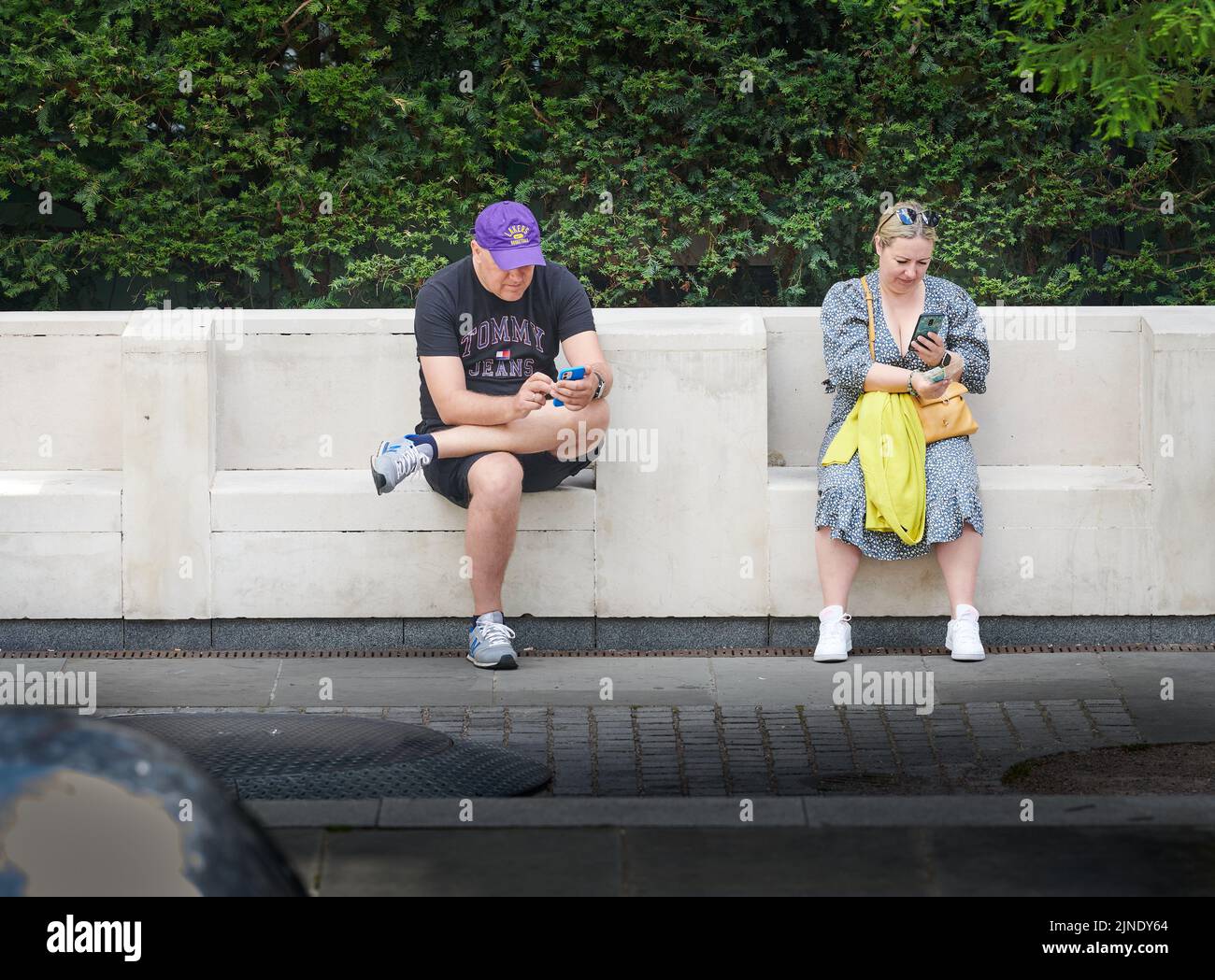Ein Duo entspannt sich, während sie auf einer Steinbank sitzen und ihre Telefone auf Peter's Hill, St. Paul's, London, England, betrachten. Stockfoto